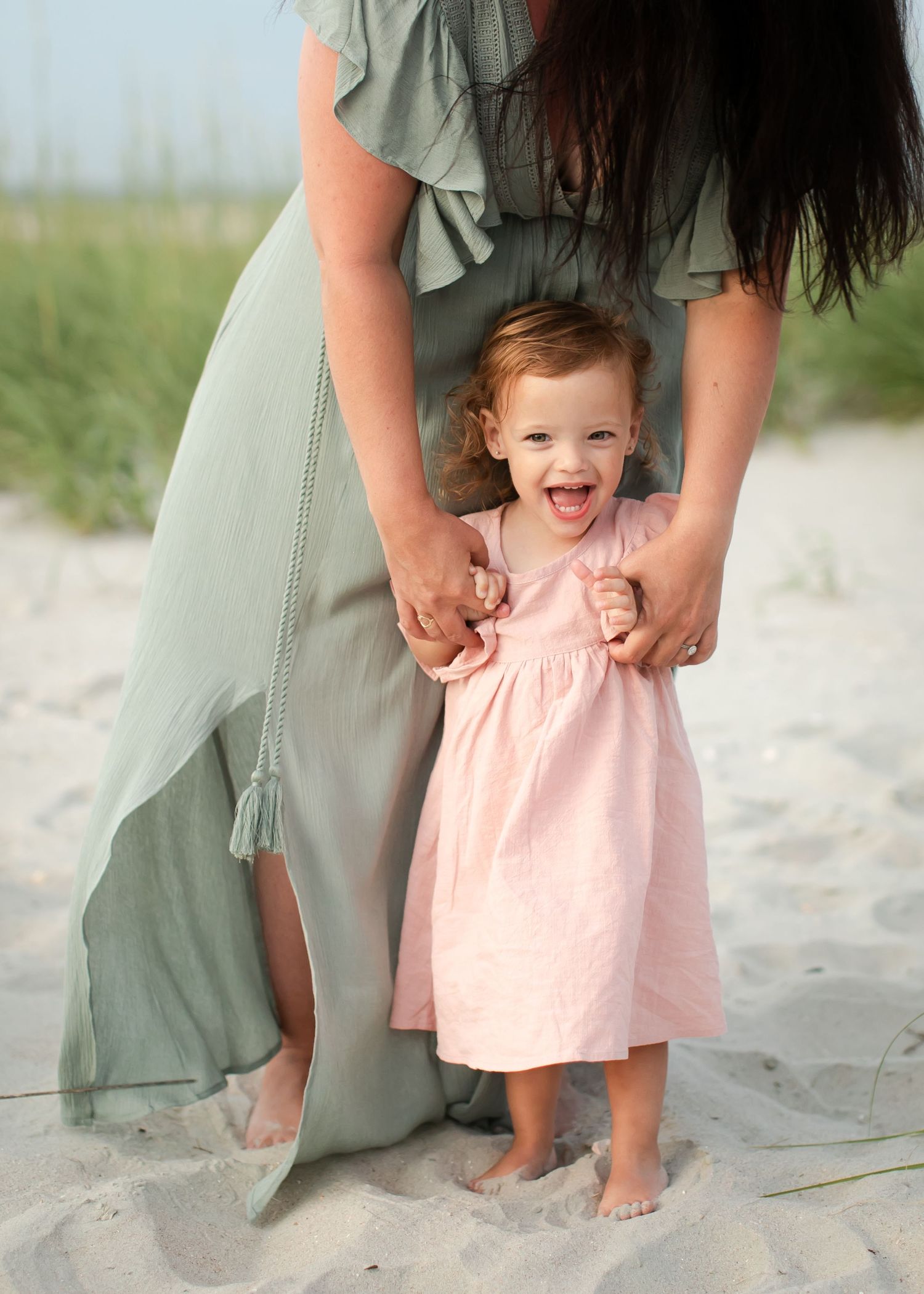 A child in a pink dress and adult in sage green dress standing together on a sandy beach.