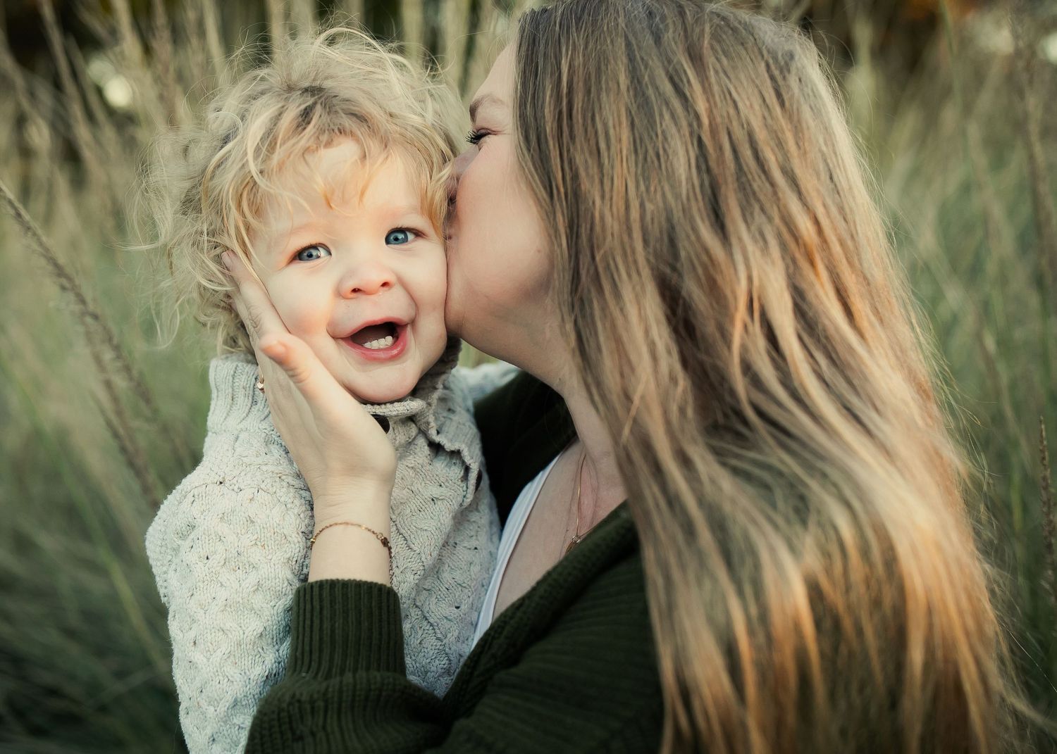 A tender moment captured outdoors as a mother gives her joyful toddler a kiss on the cheek among tall grasses.