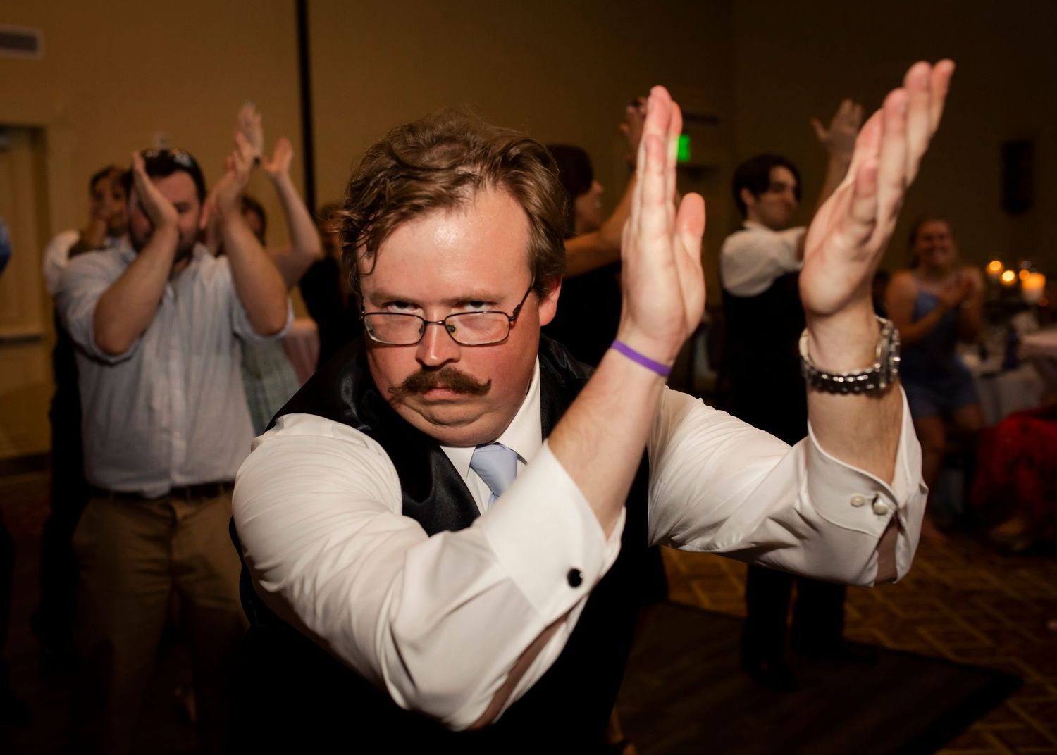 Person in glasses and white dress shirt with an amazing moustache dancing enthusiastically with raised arms at a wedding.