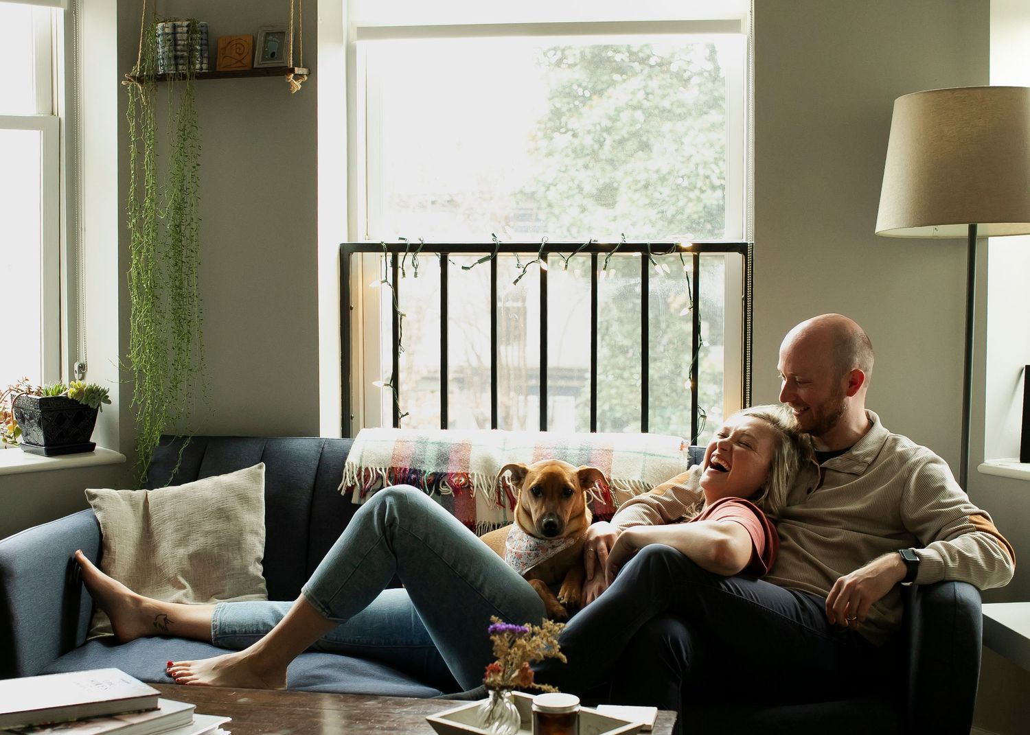 A cozy living room scene with a couple and their dog relaxing together on a couch by a bright window.