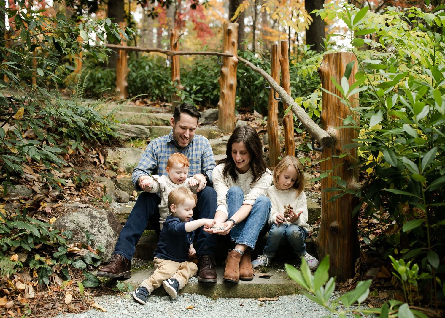 A family sits together on rustic wooden steps surrounded by lush greenery in an outdoor garden setting.
