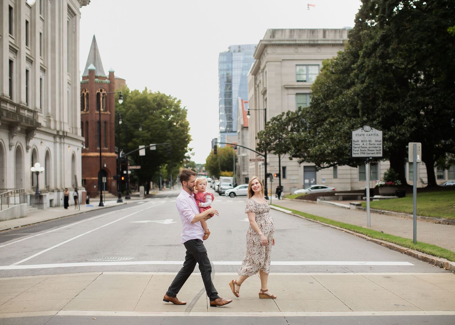 Two people and their child cross an urban street with historic buildings visible in the background.