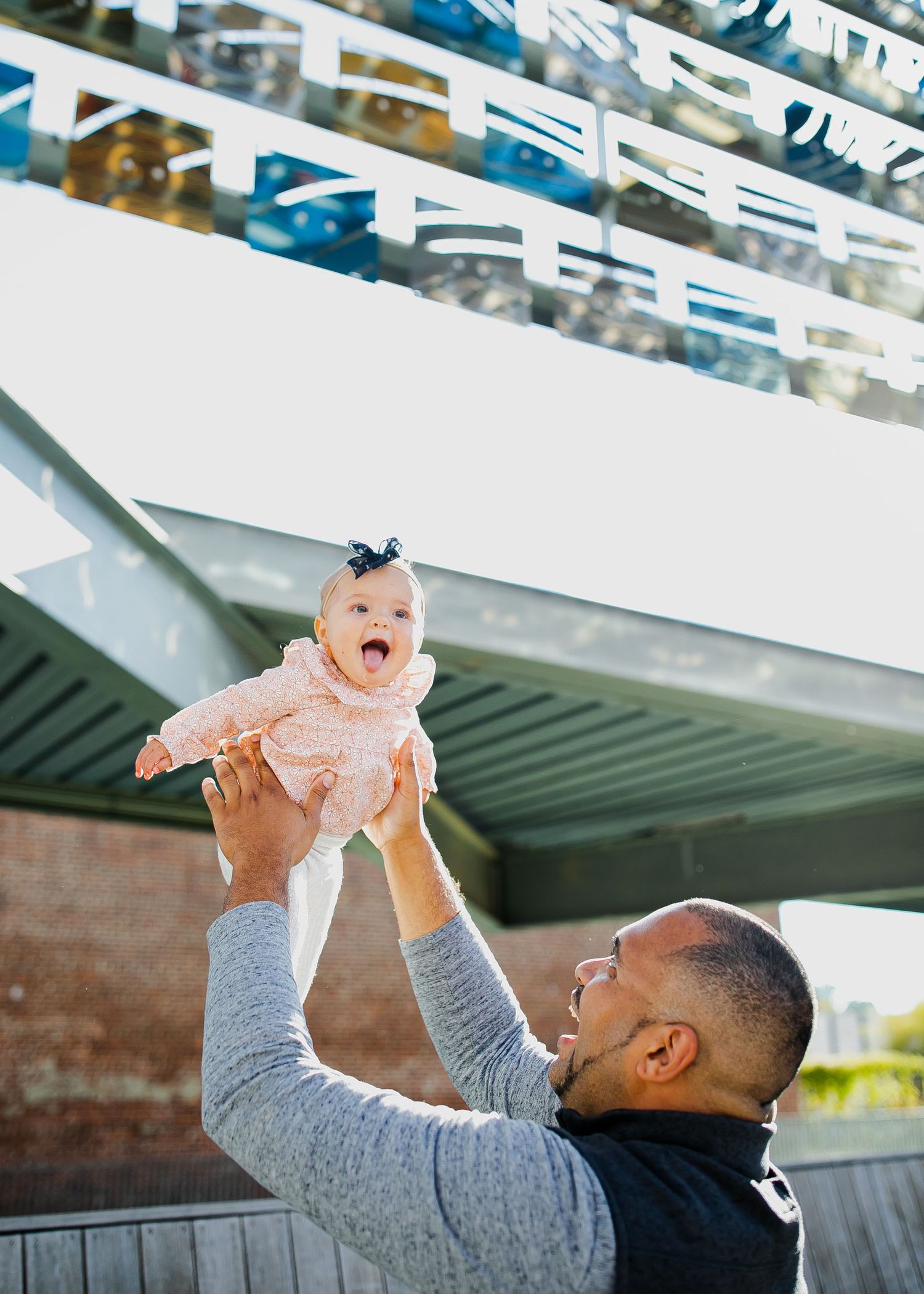 A person in a gray sweater and black vest lifts a baby wearing a pink dress overhead.