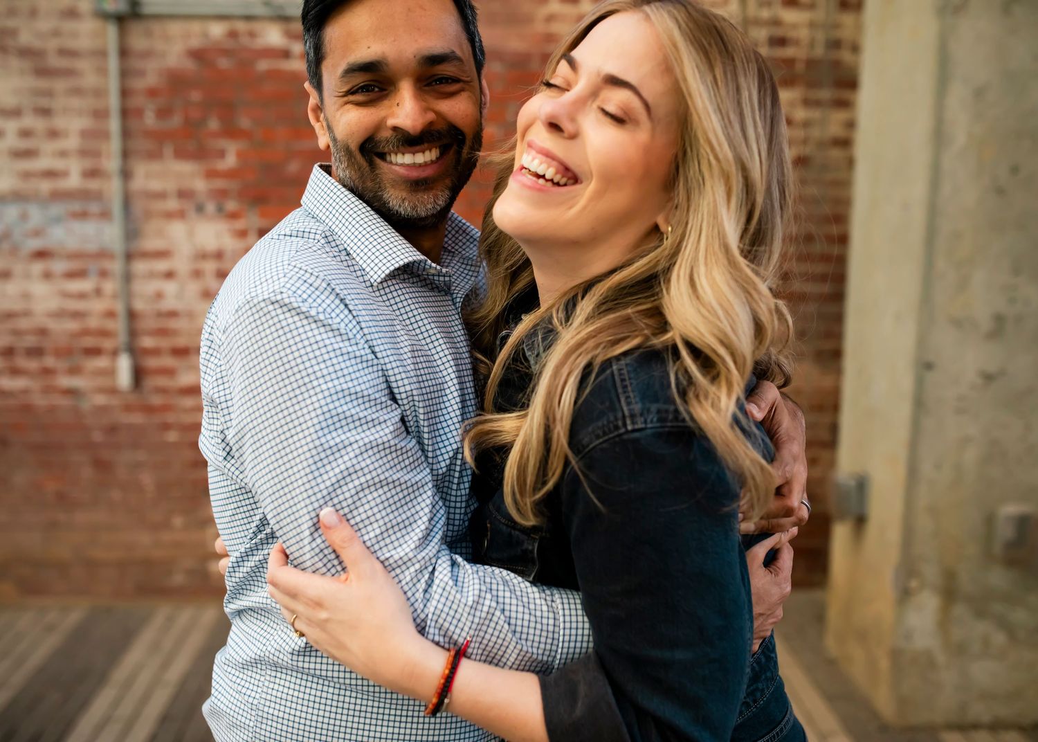 A couple laughs and embraces in front of a rustic brick wall during a romantic photoshoot.