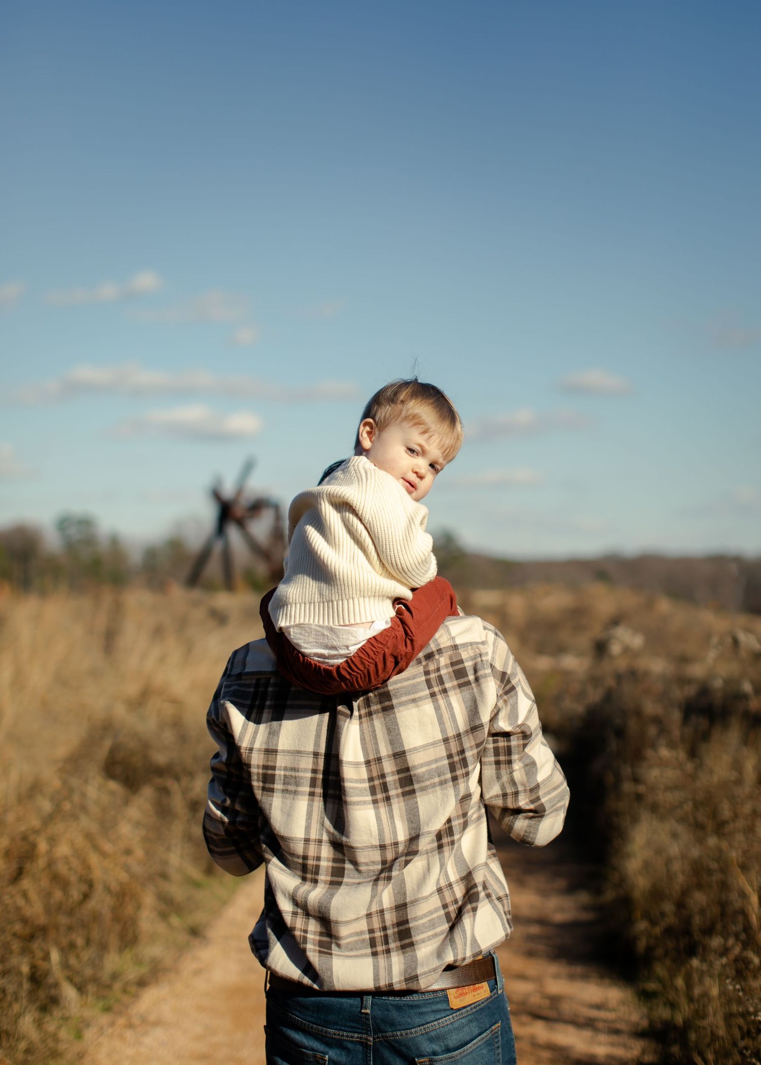 A child wearing a cream sweater is carried on someone's shoulders along a dirt path through a fall field on a sunny day.