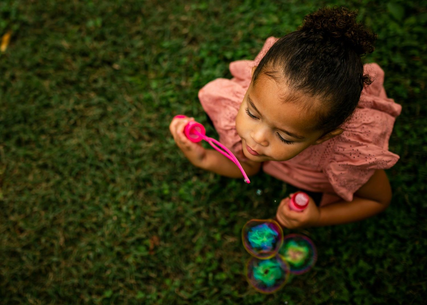 Young child in pink shirt blowing bubbles while playing outdoors on grass from aerial view.