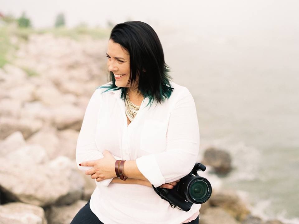 Amaris in a white shirt stands near a rocky beach shoreline on a sunny day.