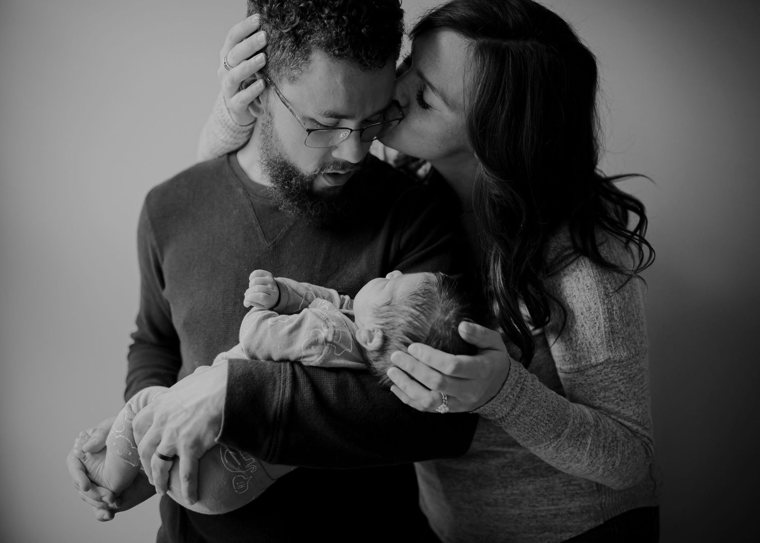 A tender black and white portrait of new parents sharing an intimate moment while holding their newborn baby.
