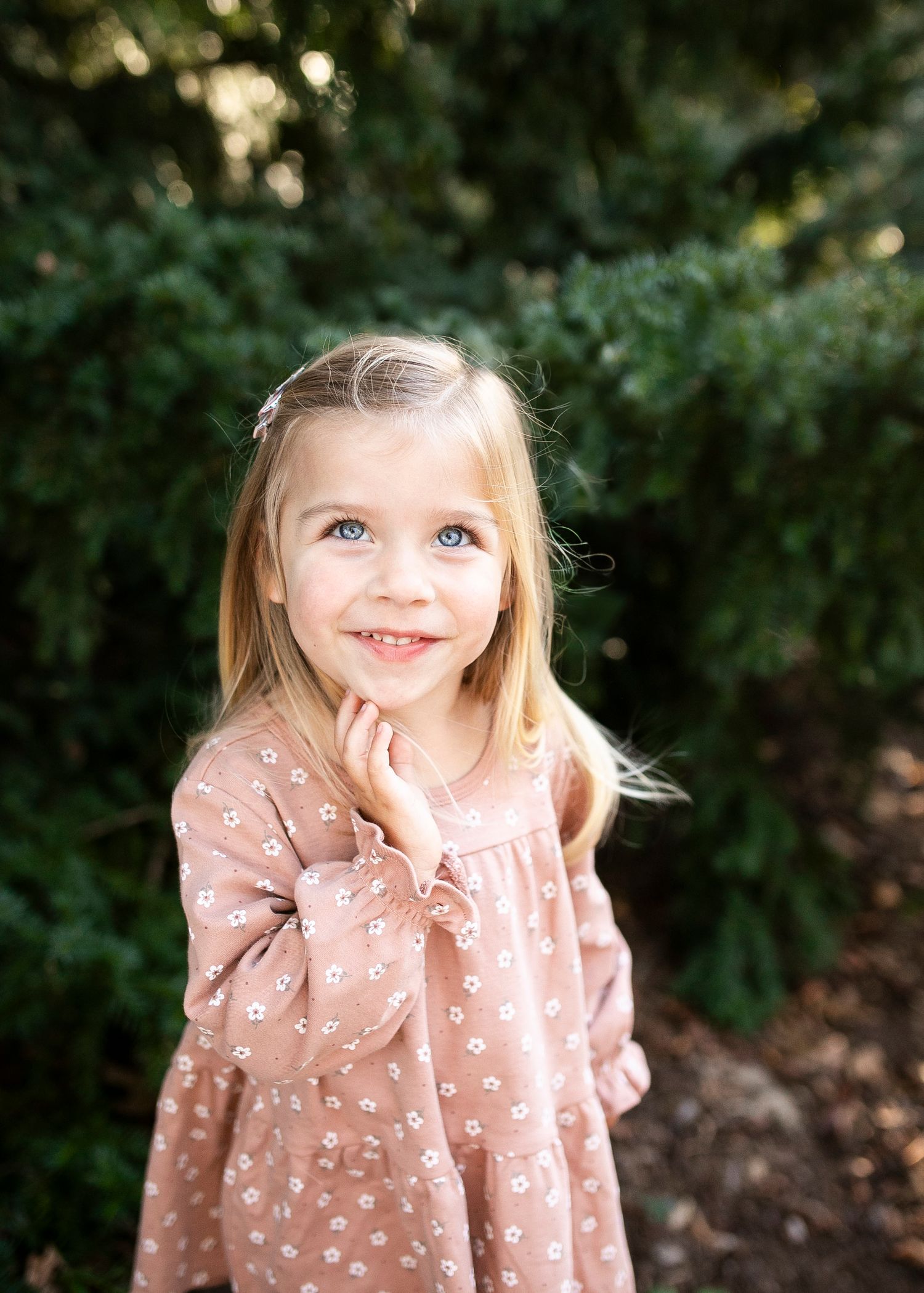 A young child in a dusty pink ruffled dress smiles and poses against a blurred green outdoor background.