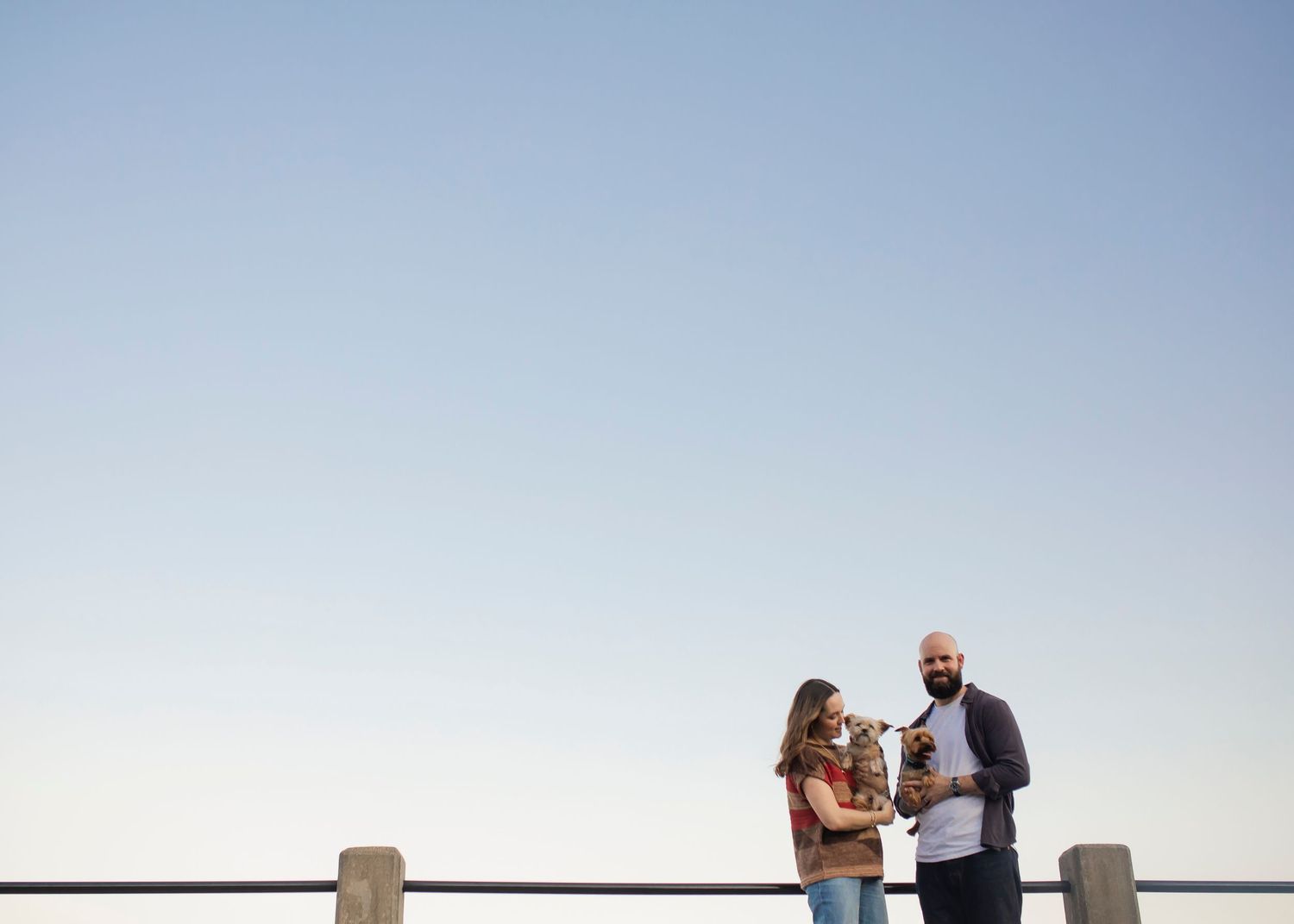 Two people stand together holding their pets against a minimalist backdrop of clear sky and a simple railing.