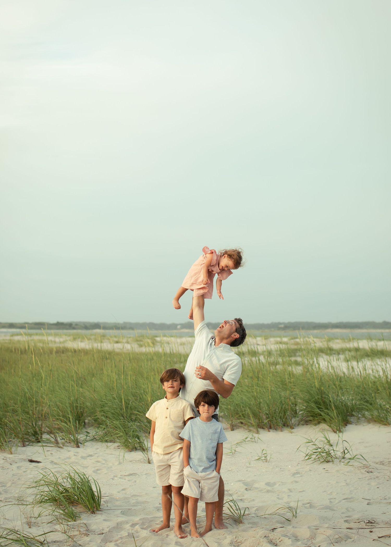 Family enjoying a playful moment together on a sandy beach with sea grass and ocean in the background.