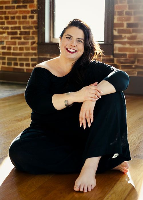 Photo of Amaris sitting on a wooden floor in front of a brick wall with a bright window. Amaris is barefoot.