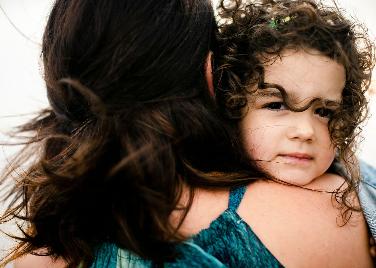 Mother holding a child. She is facing away and the child faces the camera. The wind has blown a lock of curly hair across the child's face.