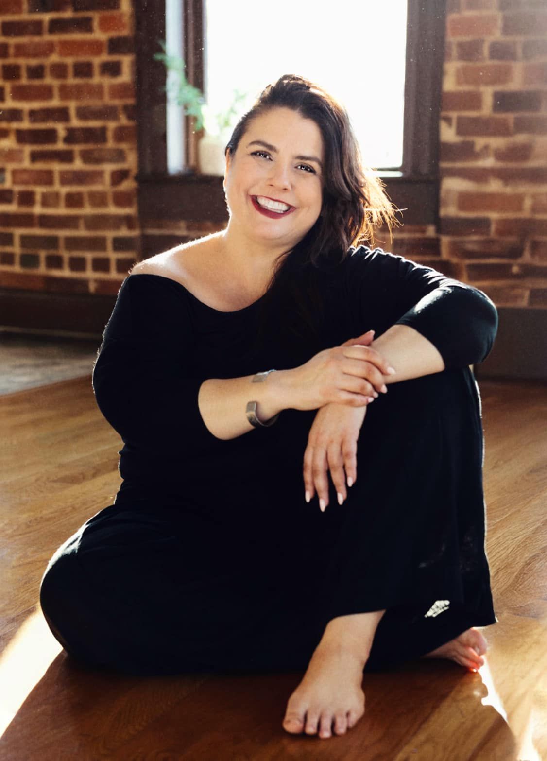 Amaris poses casually on a hardwood floor against exposed brick walls in an industrial space.