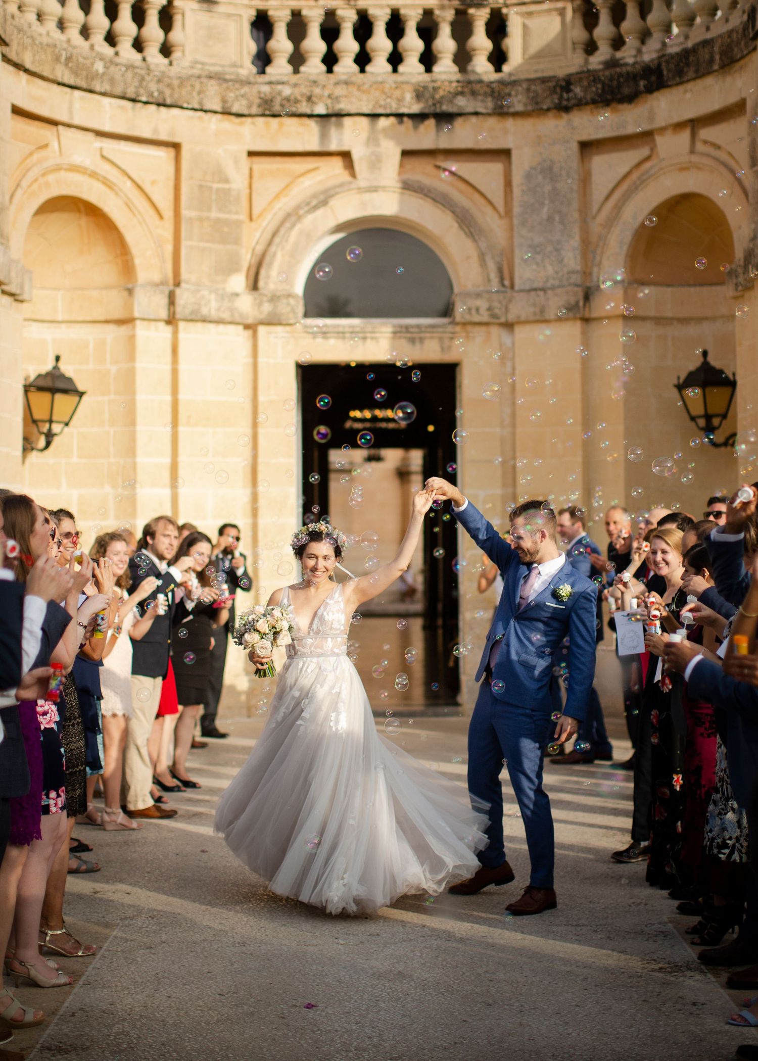 A view of a wedding couple dancing with guests in a grand stone courtyard at dusk.