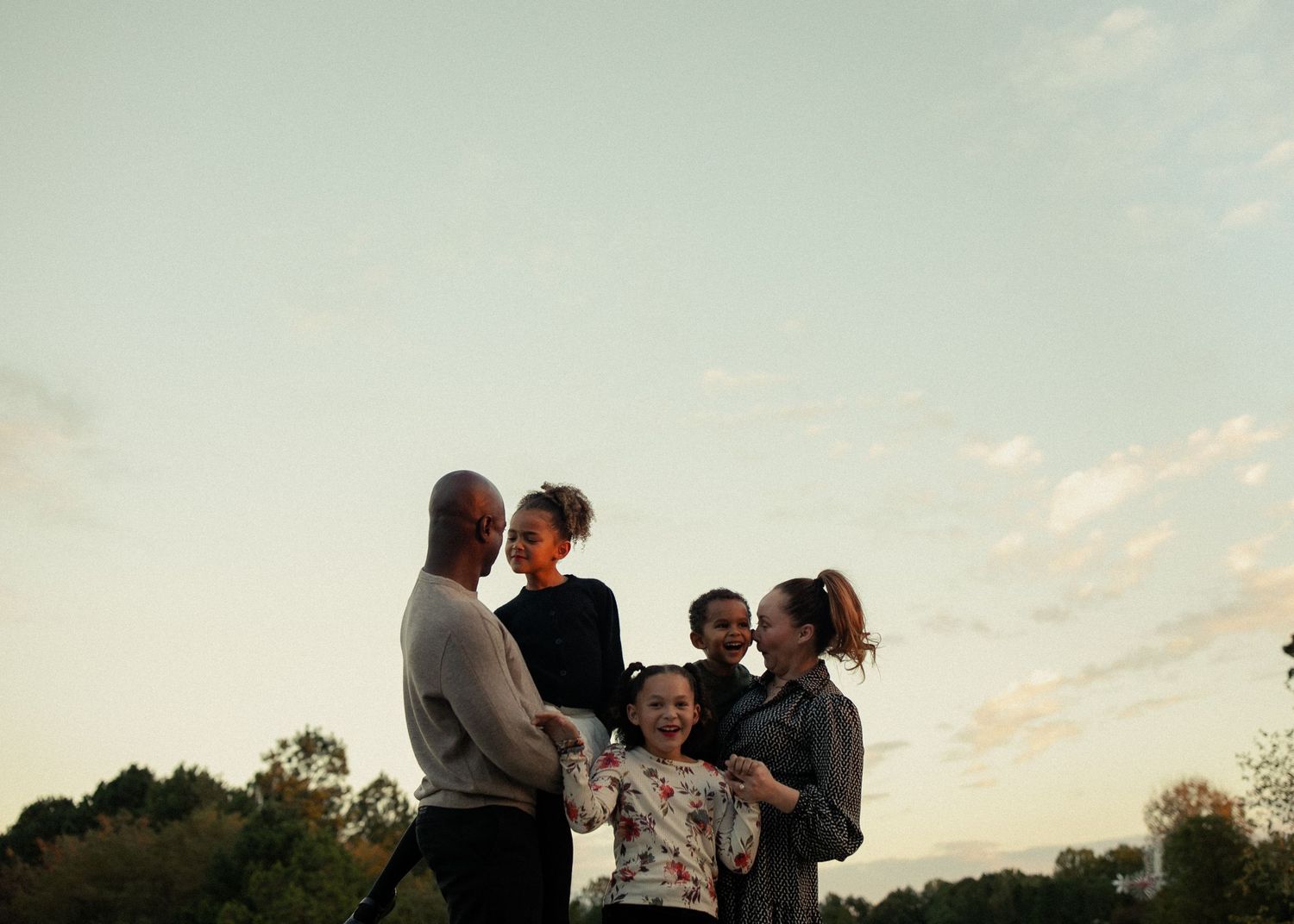A group of people gather outdoors during golden hour, silhouetted against a soft sunset sky.
