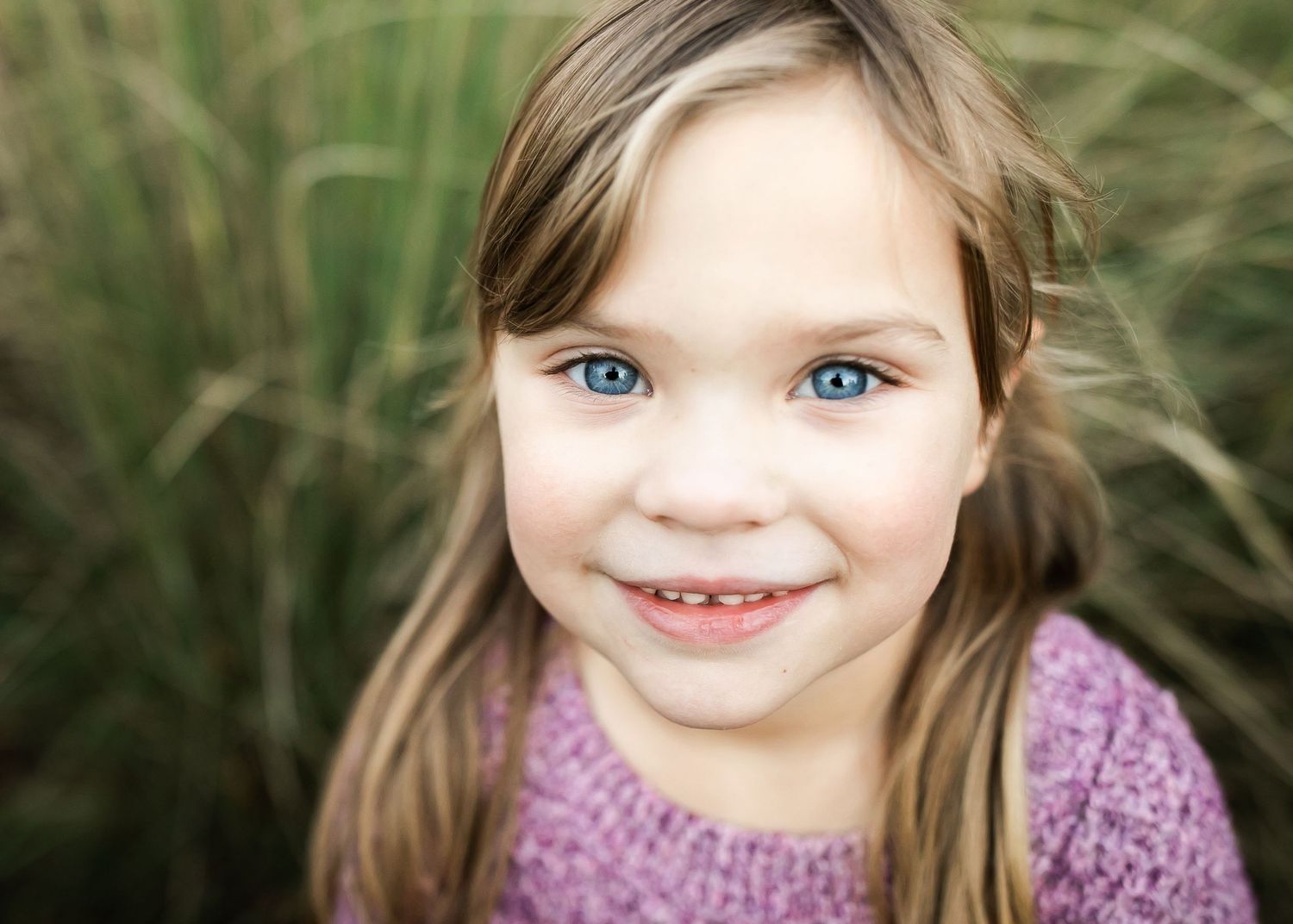 A bright-eyed child wearing a purple dress smiles warmly against a backdrop of tall grass.