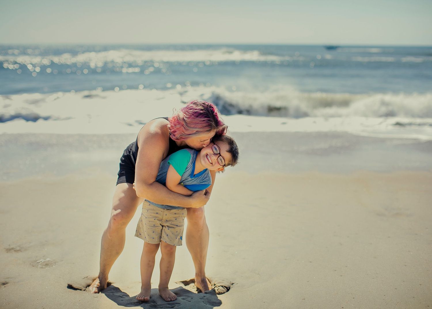 A woman embraces a child as they laugh together on a sunny beach with ocean waves in the background.