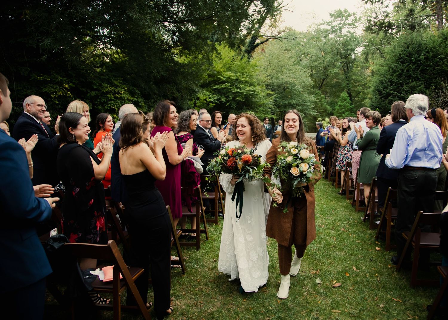 Wedding guests gather outdoors in a garden setting during a summer wedding ceremony. The couple walks down the aisle with their bouquets.