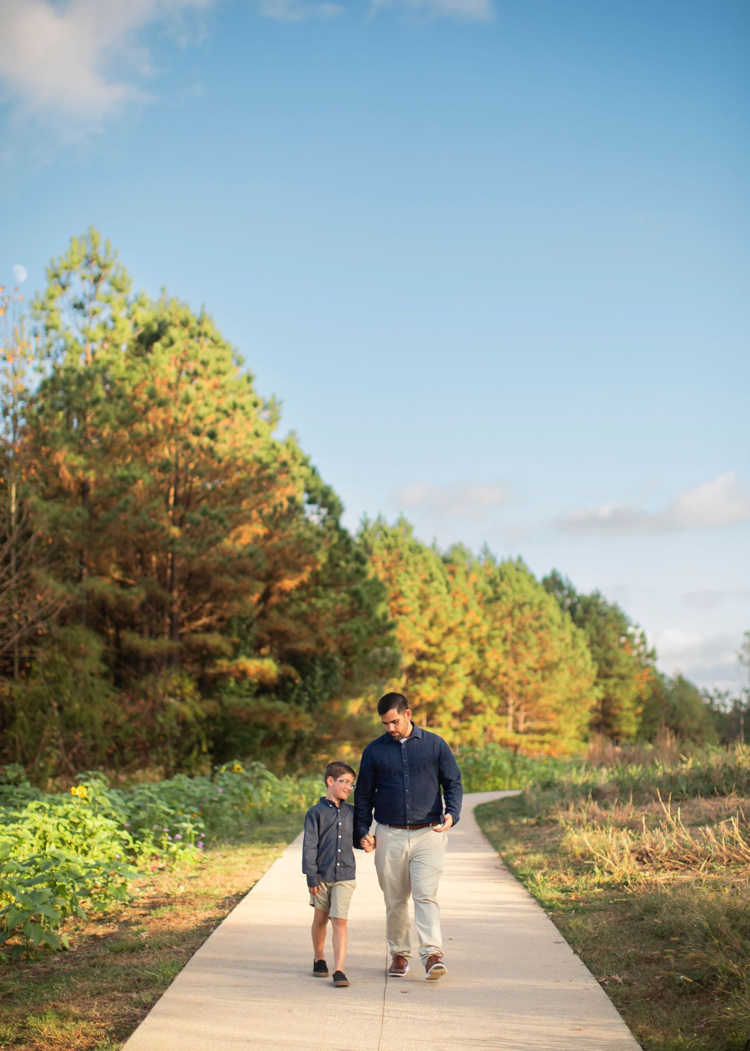 Two people walk together on a wooden boardwalk path surrounded by autumn trees and greenery on a sunny day.