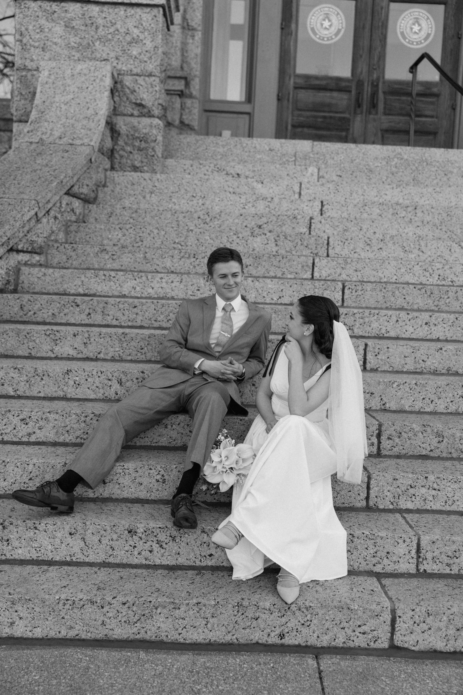Bride and groom sit together on stone steps in black and white wedding portrait.