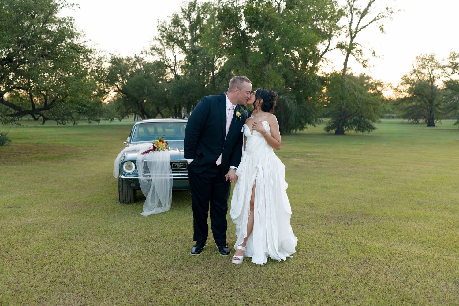 Bride and groom sharing a romantic kiss in front of a vintage car on a lush green lawn at golden hour.