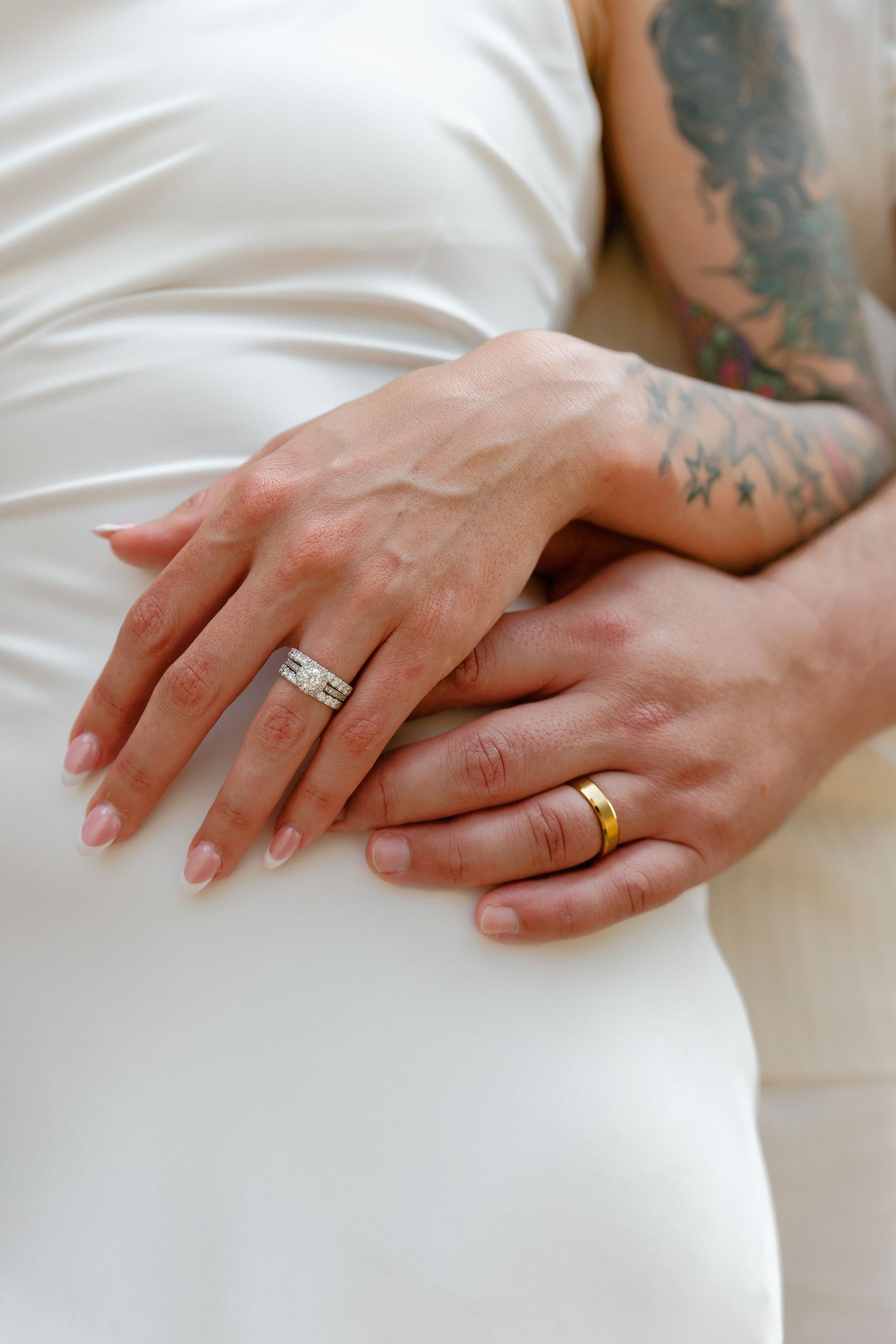 Close-up view of hands wearing wedding rings resting on white fabric showing tattoo details and engagement ring.