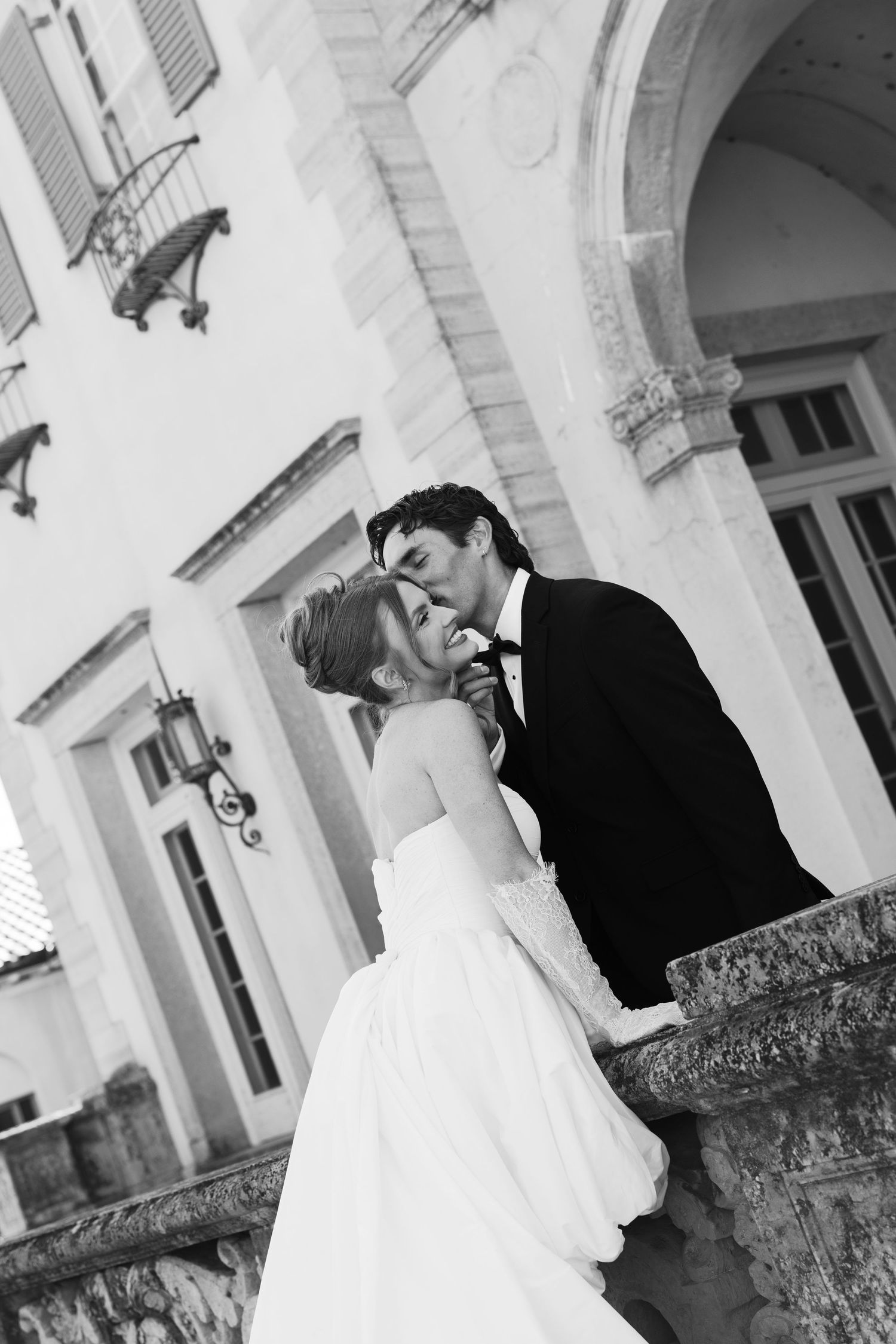 Black and white photo of a bride and groom sharing a kiss on elegant stone steps outside a classic mansion.