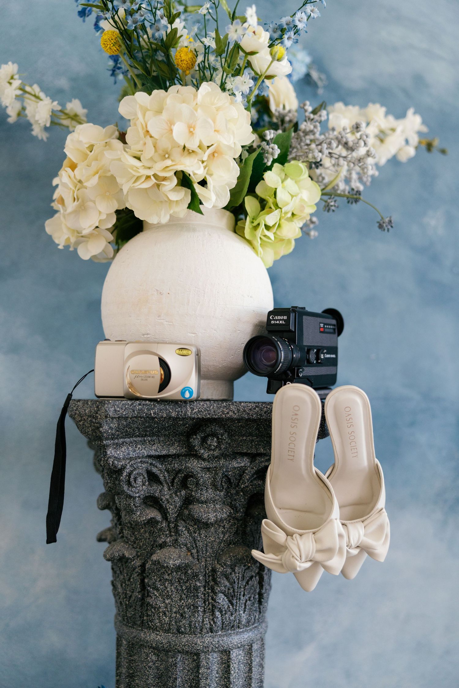 White floral arrangement in a cream vase sits atop a textured gray stone pedestal against a cloudy blue backdrop.