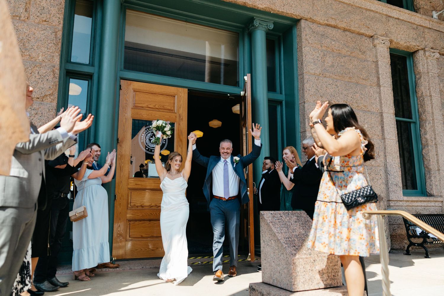 Joyful celebration outside historic building entrance with group throwing confetti and cheering in bright sunlight.