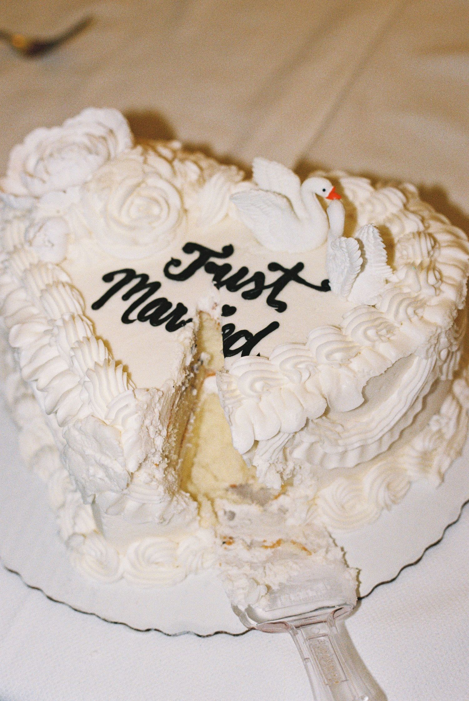 White wedding cake decorated with roses and swans, reading Just Married, with a slice being served.