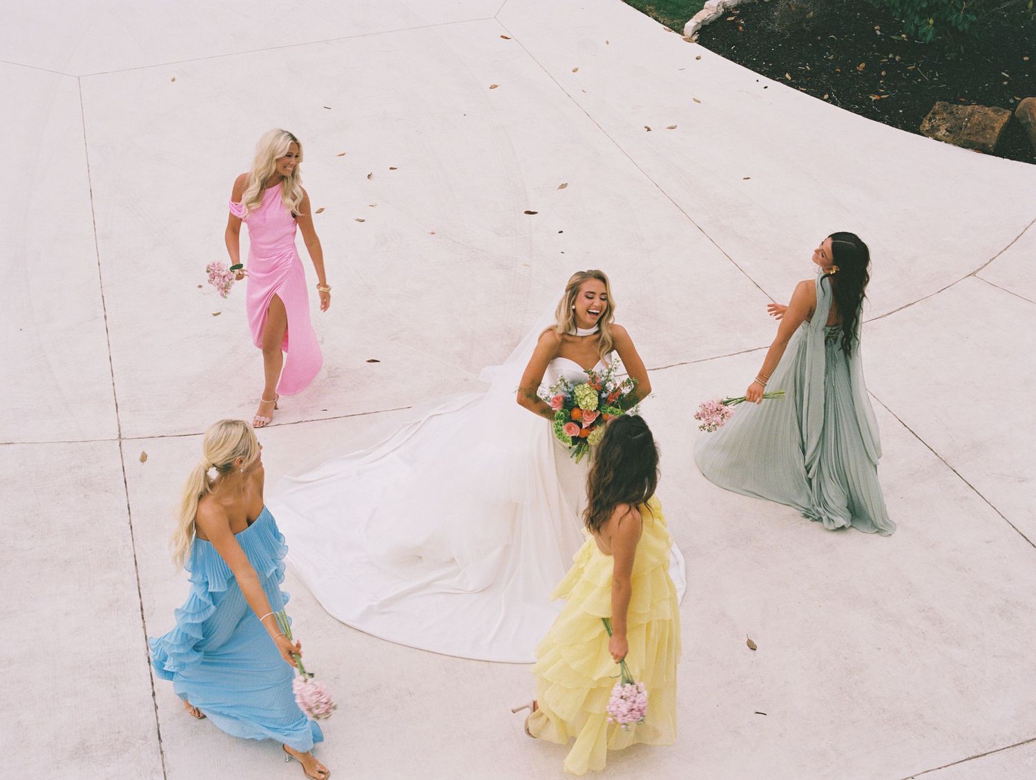 Bride in white gown surrounded by bridesmaids in pastel dresses outdoors from aerial view.