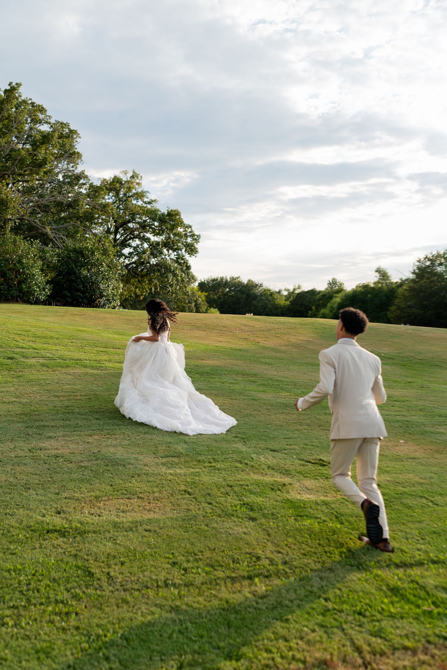 A couple in wedding attire runs playfully across a green hillside on a cloudy afternoon.
