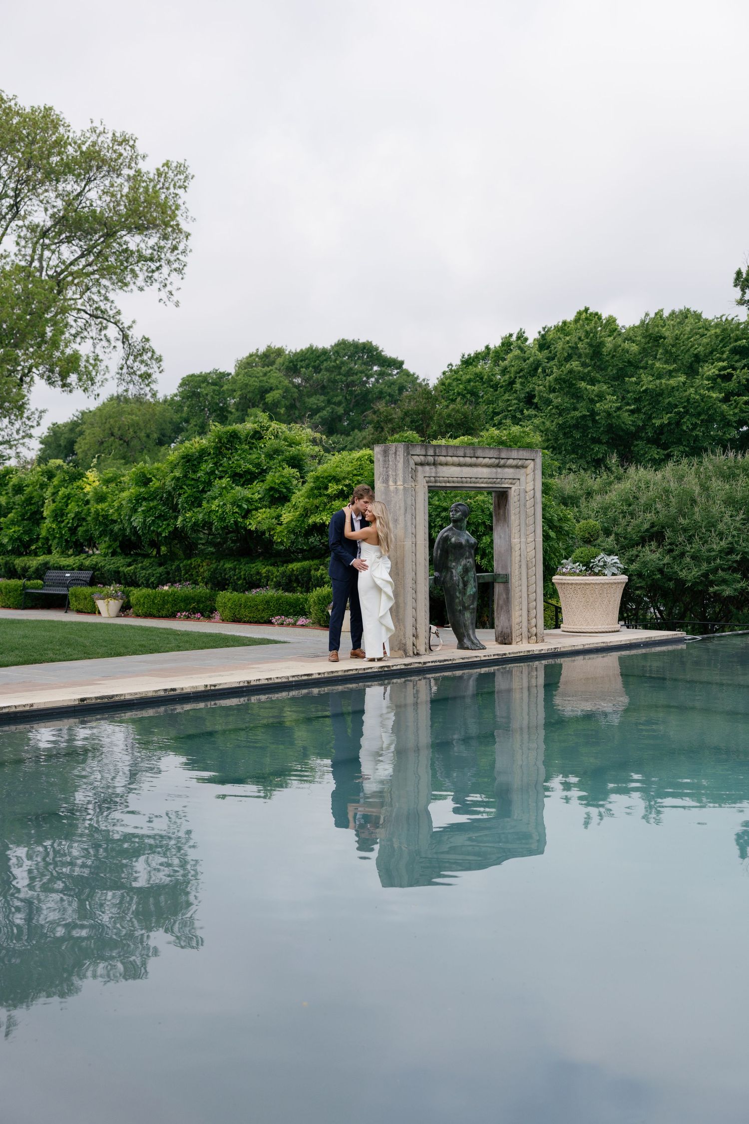 A bride and groom embrace by a reflective pool beside a stone arch sculpture in a lush garden setting.