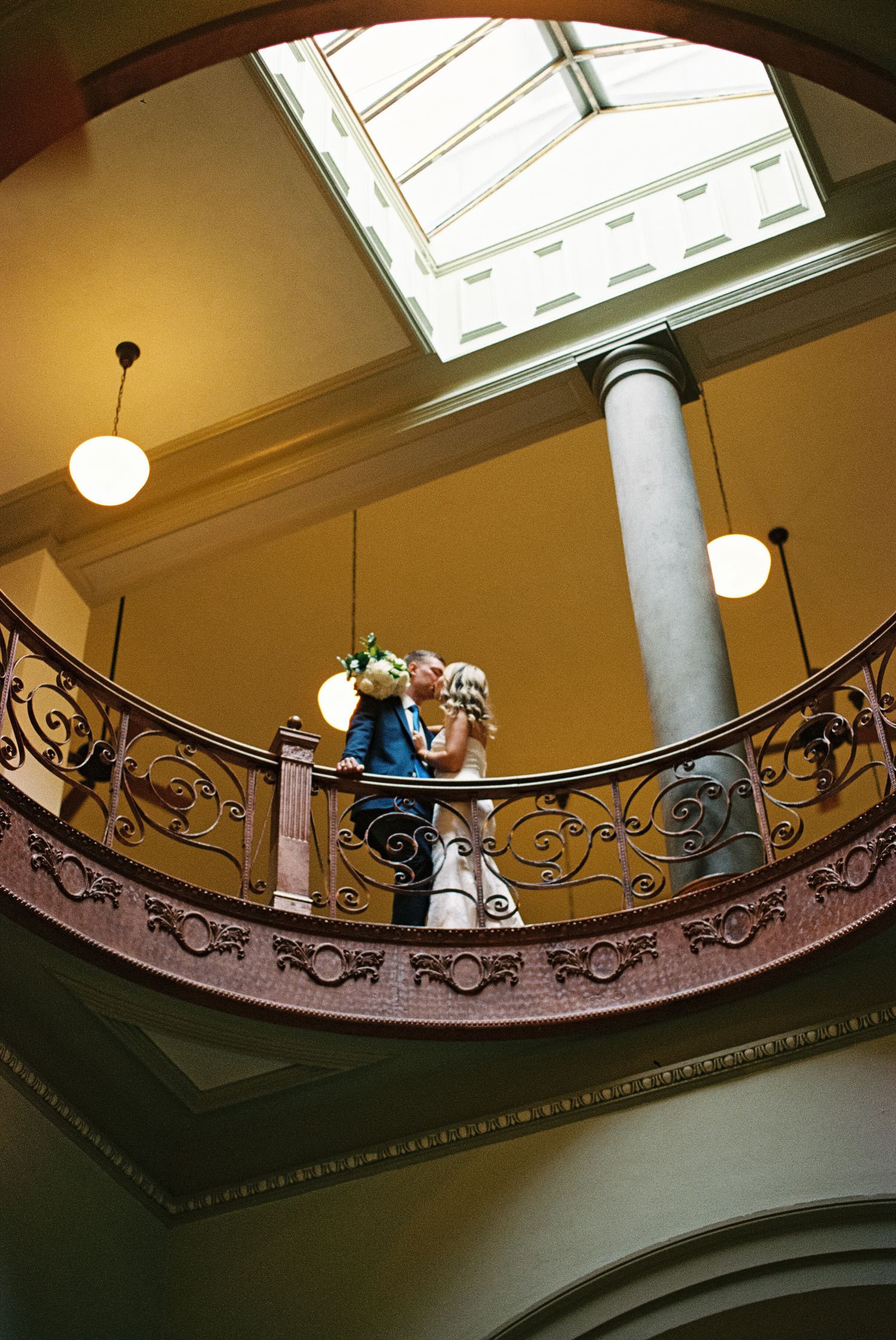 Couple sharing a romantic kiss on an ornate iron balcony inside an elegant venue with a skylight.