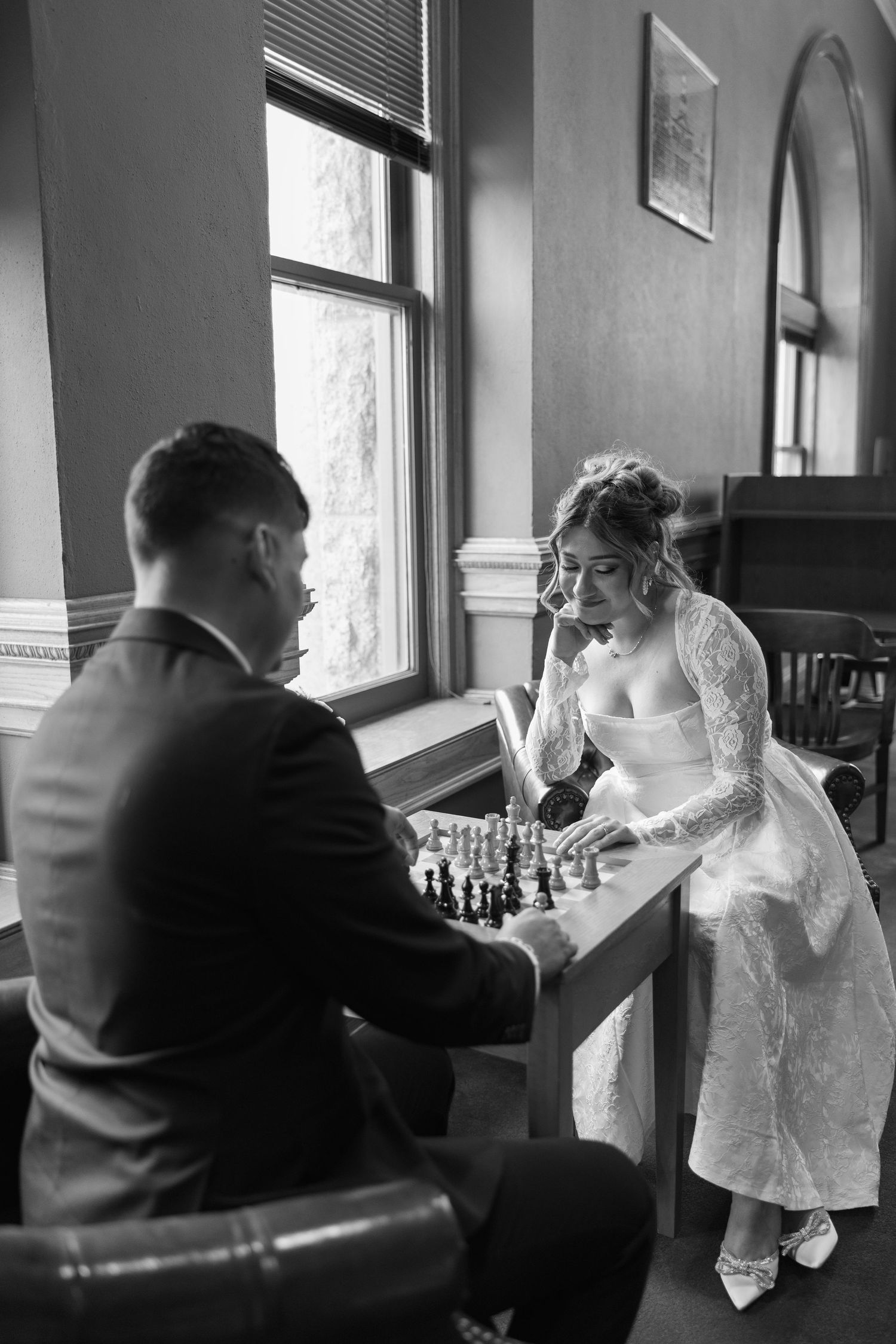Black and white photo of bride and groom playing chess by window on wedding day.