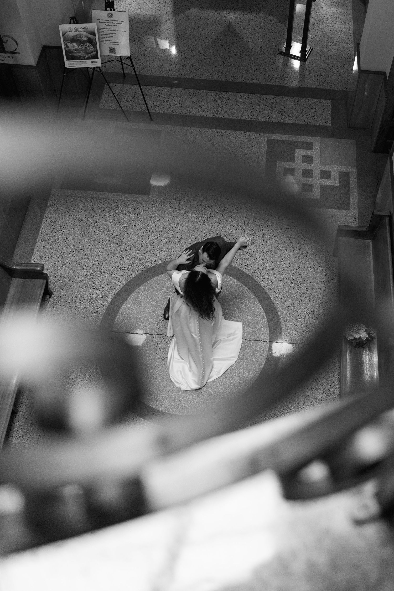 Aerial black and white photo of a woman in a white dress standing in a circular floor pattern inside a building.