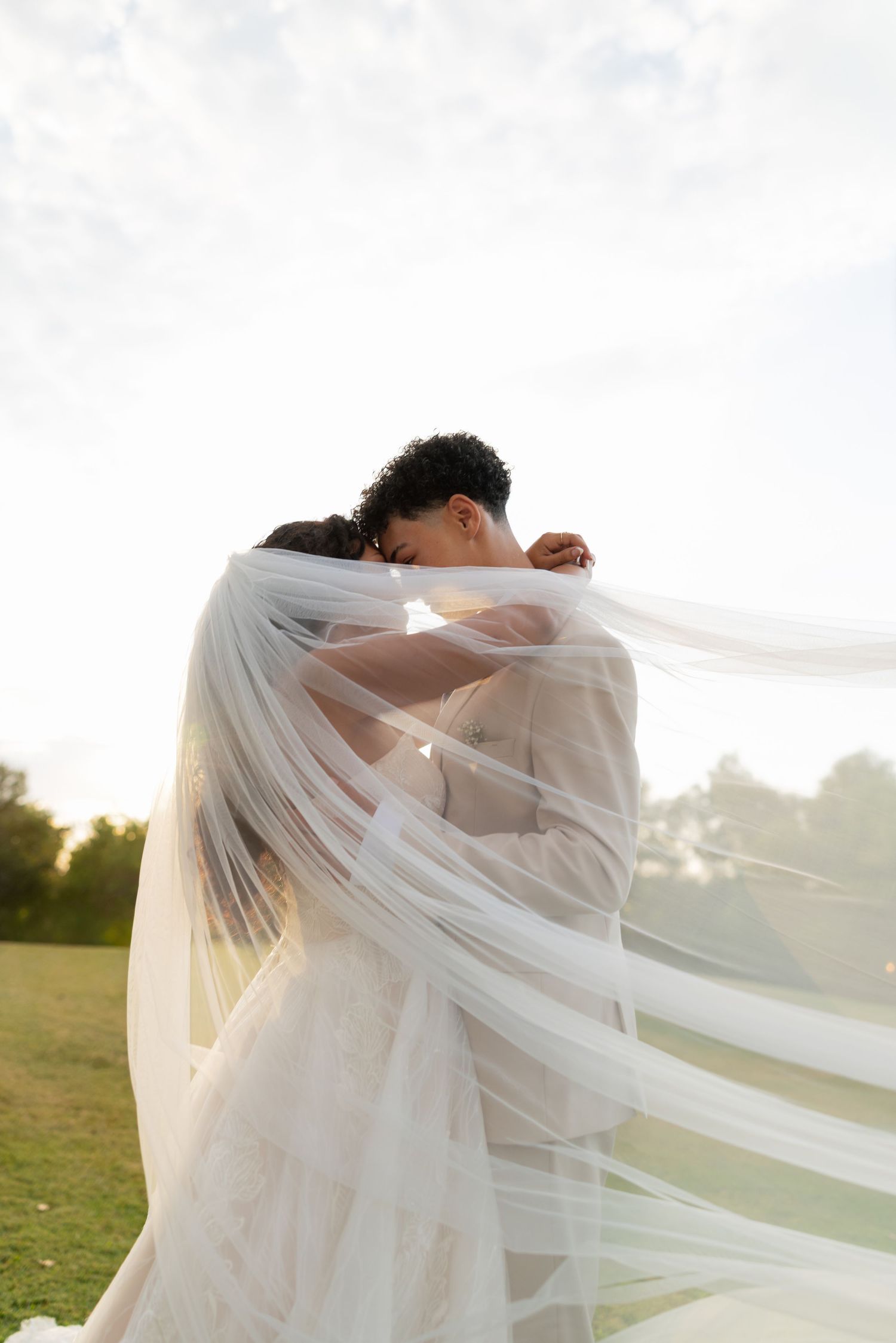A romantic wedding couple embraces under a flowing white veil during a sunset photoshoot in a grassy field.