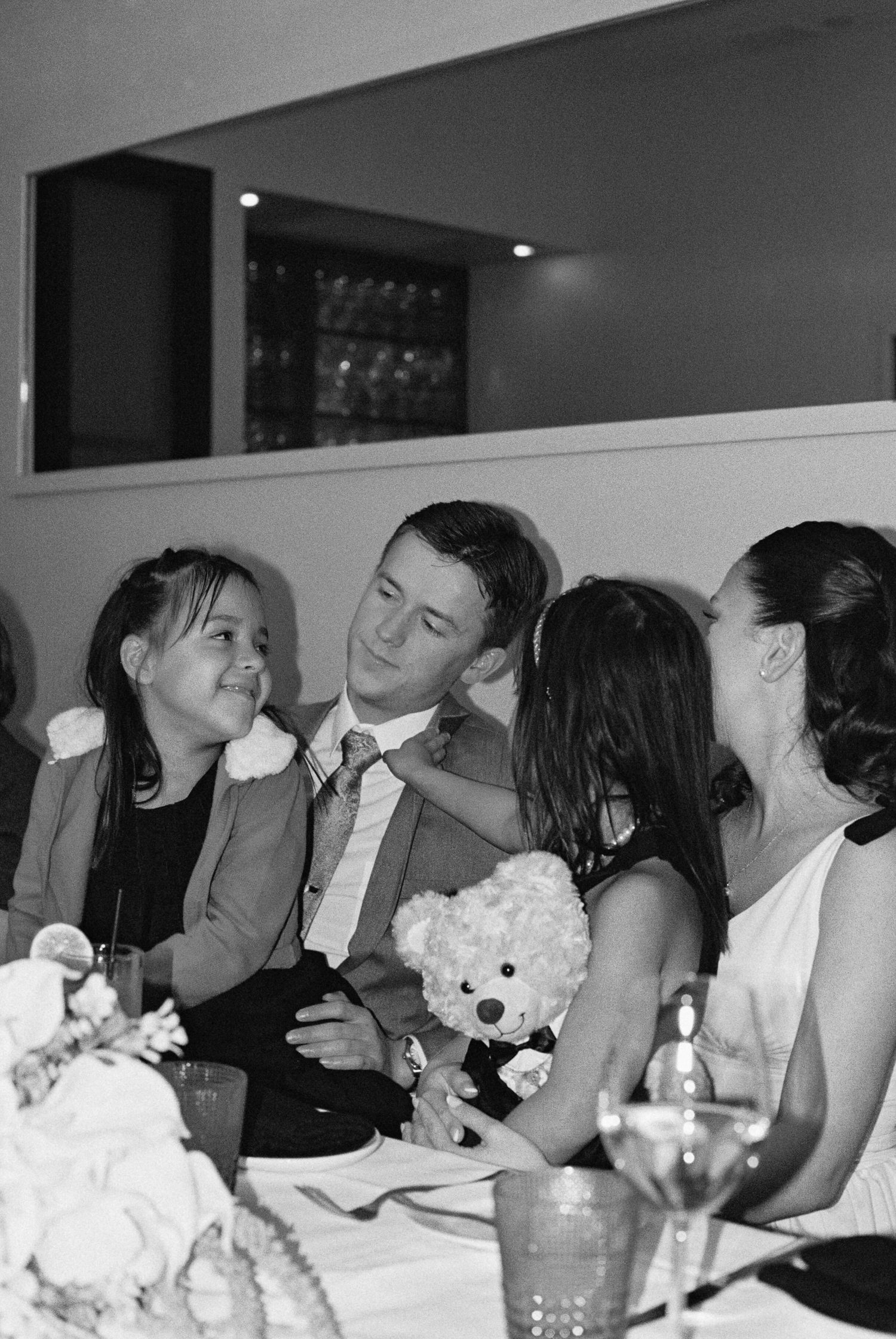 Groom sits between two young girls holding a teddy bear at wedding reception table.
