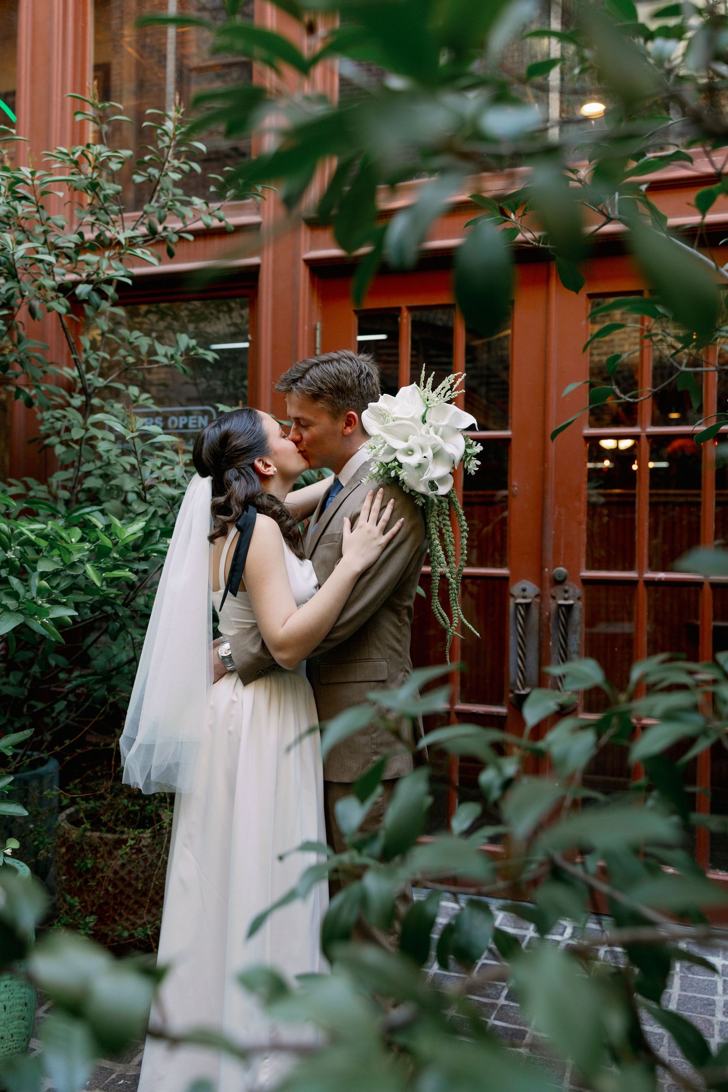 Bride and groom share a romantic kiss surrounded by lush greenery outside a rustic wooden building.