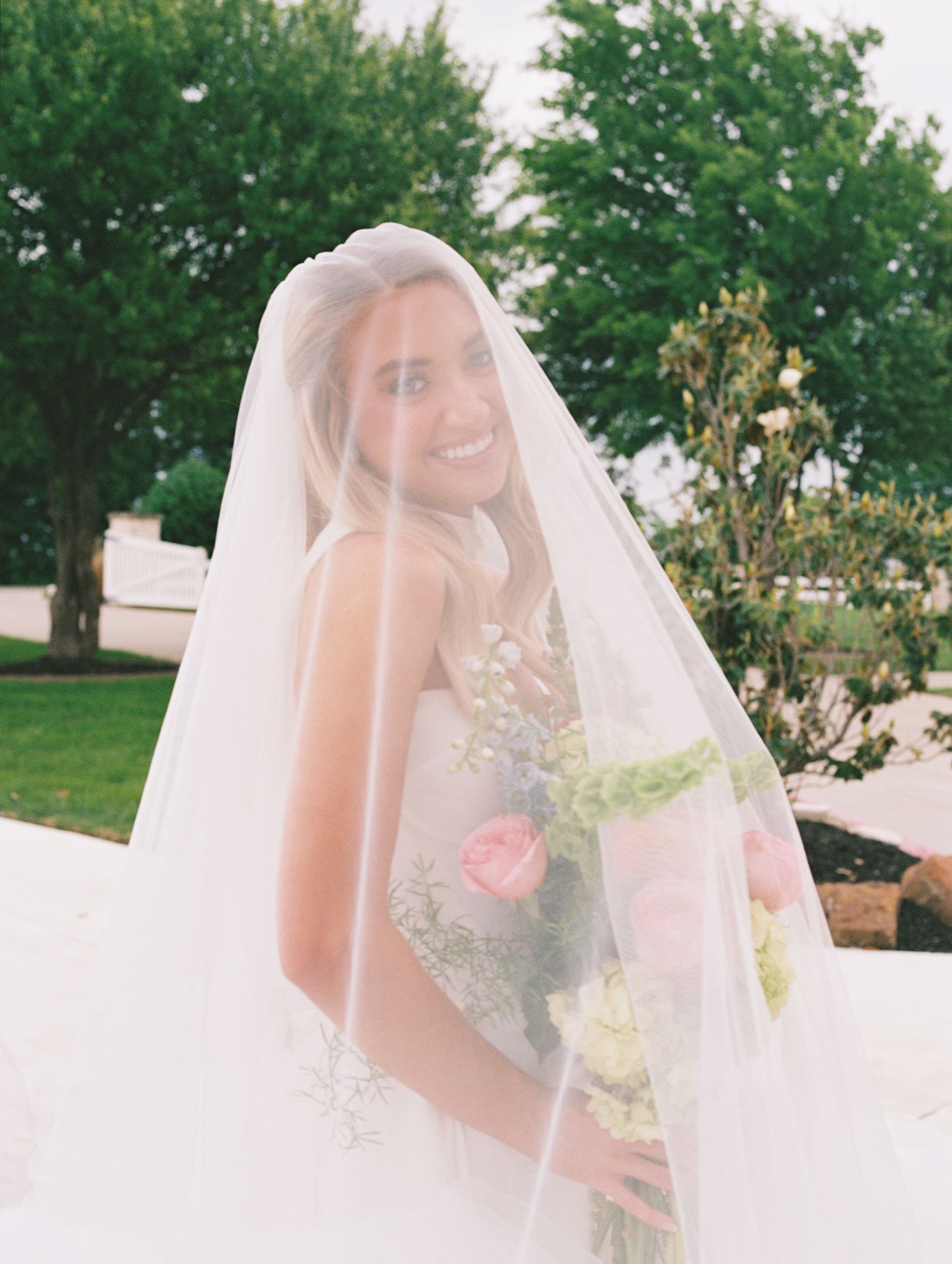 Smiling bride wearing a flowing white veil and holding a bouquet of pink and white flowers outdoors.