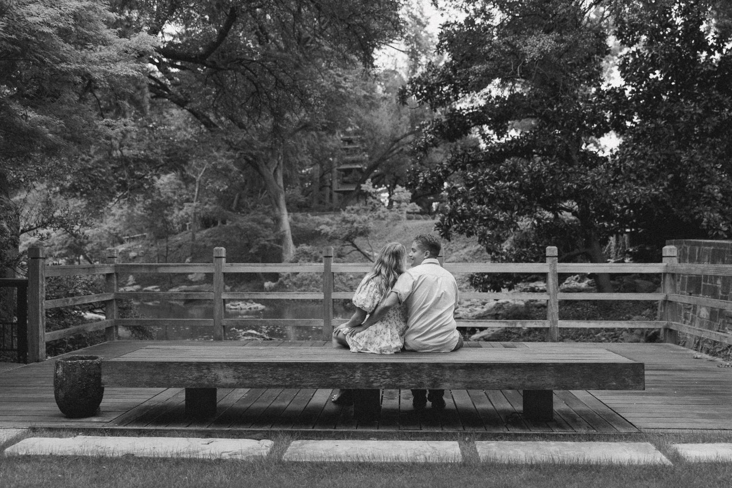 A couple sits together on a wooden bench in a peaceful garden setting, captured in black and white photography.