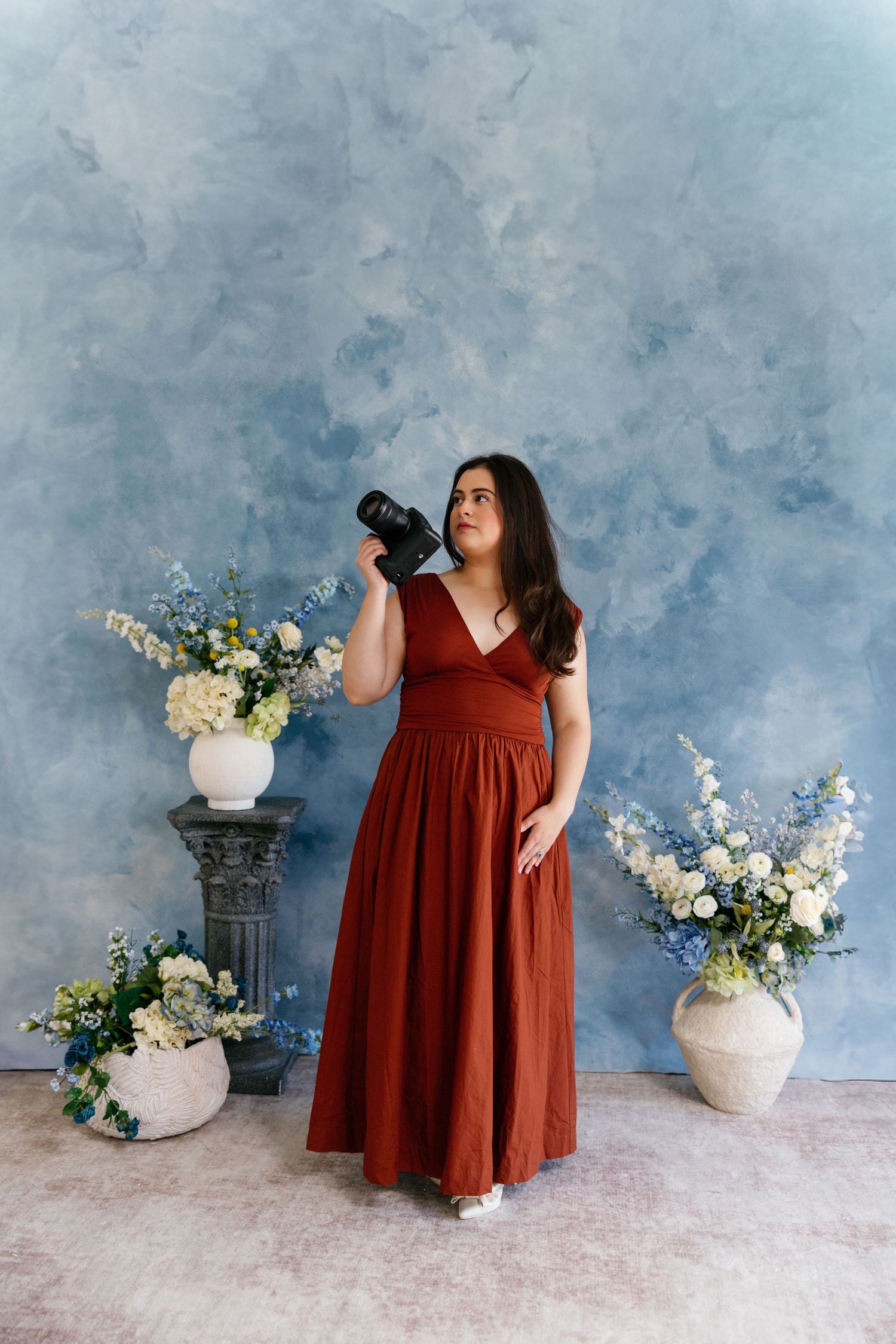 Woman in burgundy dress poses gracefully against blue backdrop with white floral arrangements.