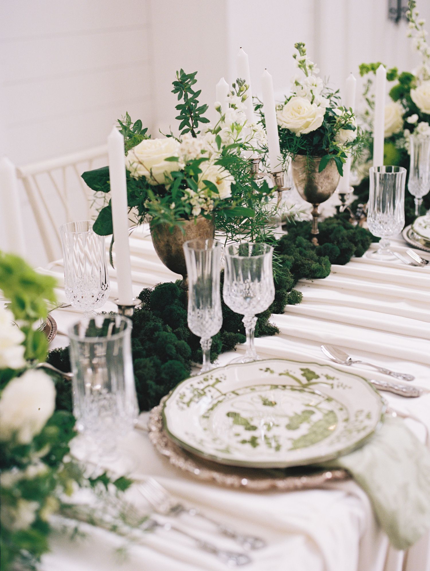 Elegant wedding table setting with white florals, greenery runner, crystal glasses, silver chargers, and white taper candles.
