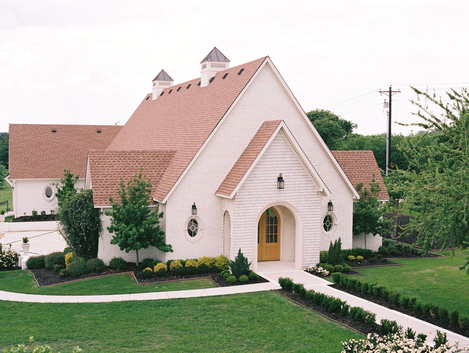 White brick chapel with arched wooden door, red roof, manicured landscaping, and tall trees on a cloudy day.