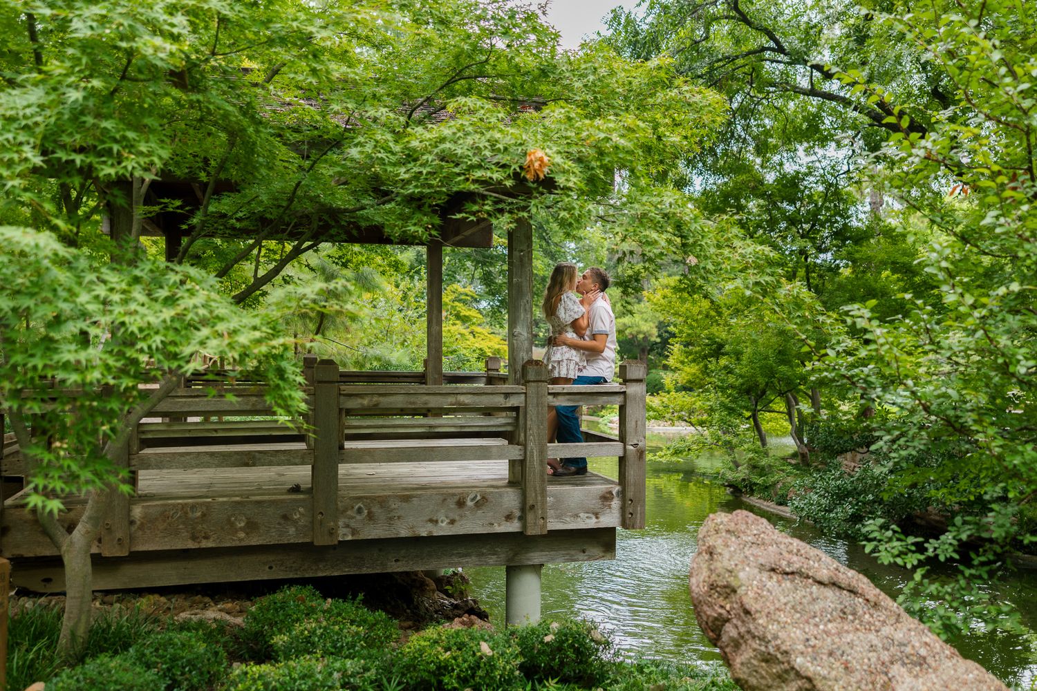 A wooden footbridge stretches across a scenic river surrounded by lush green foliage in a peaceful park setting.
