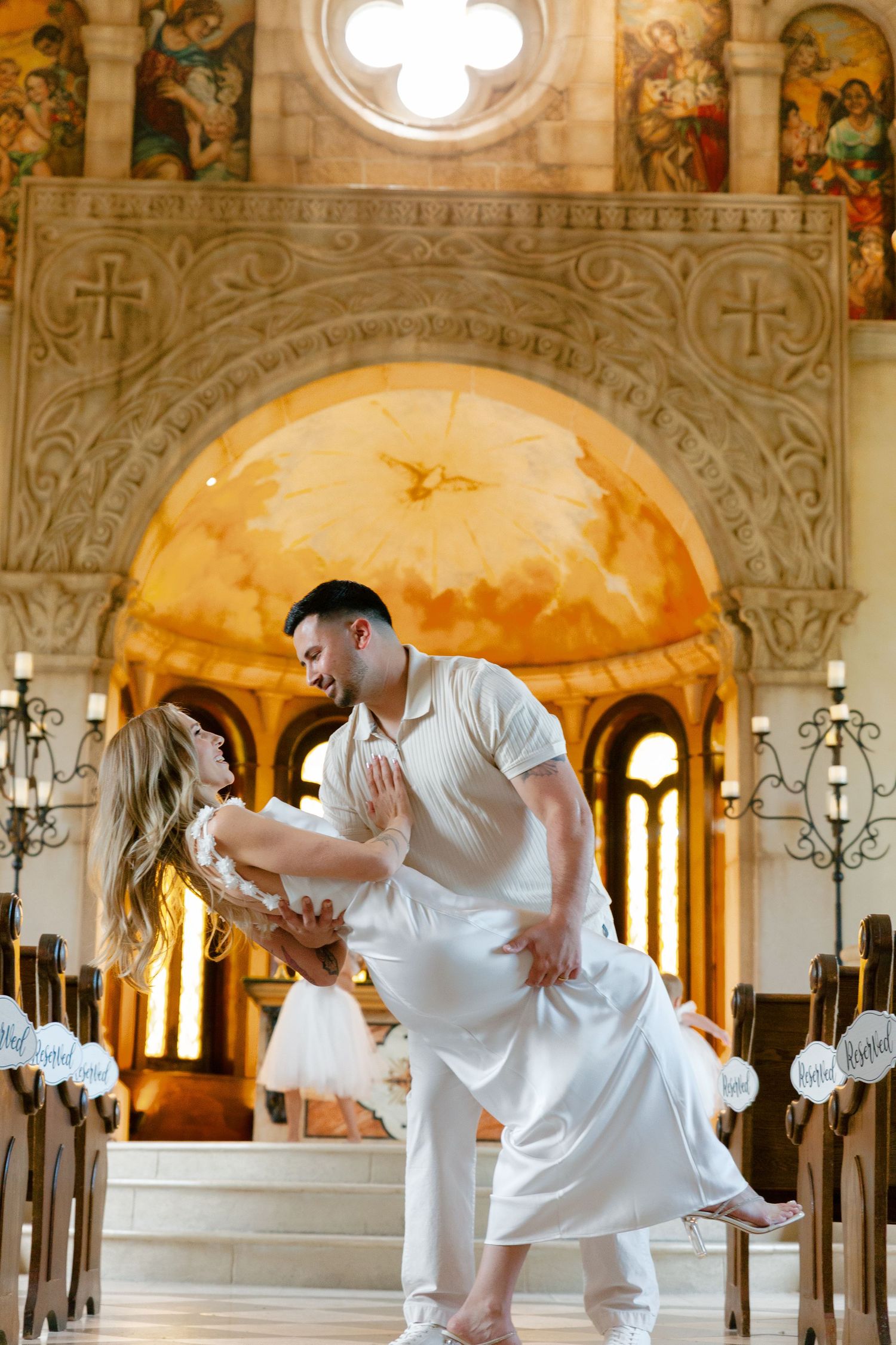 A couple in white outfits perform a contemporary dance routine inside an ornate church with vaulted ceilings and arches.