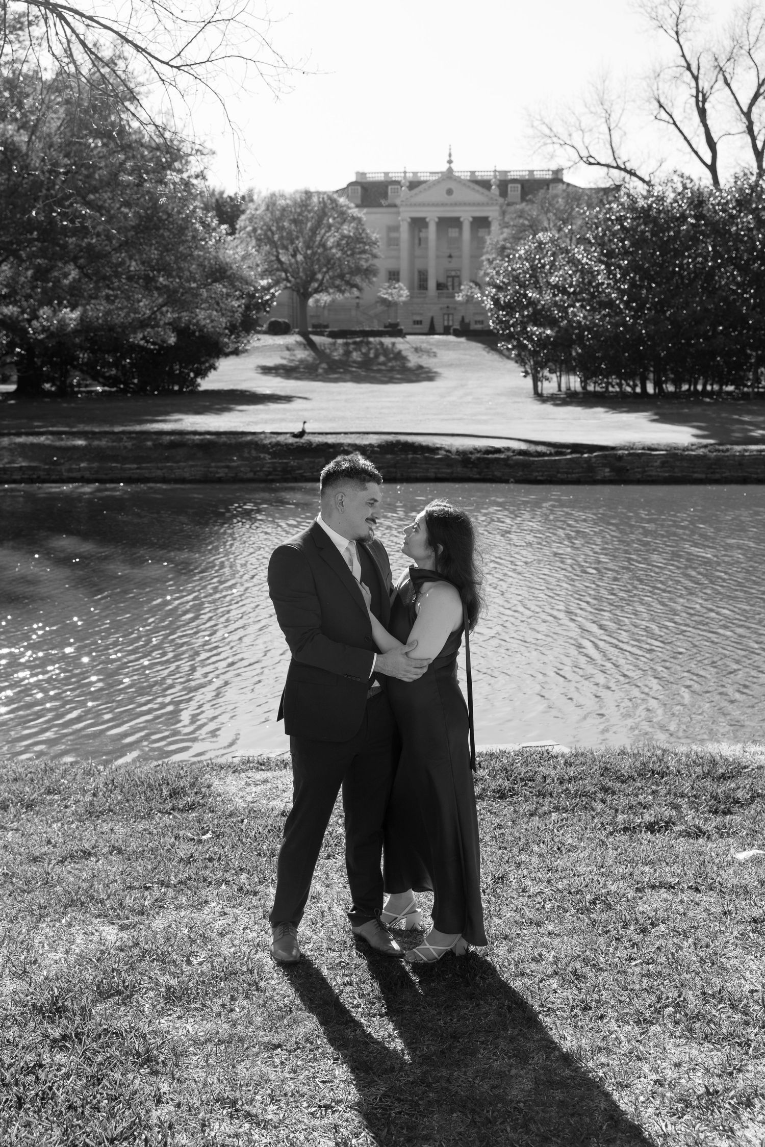 Black and white photo of a couple embracing by a pond with a grand estate building in the background.