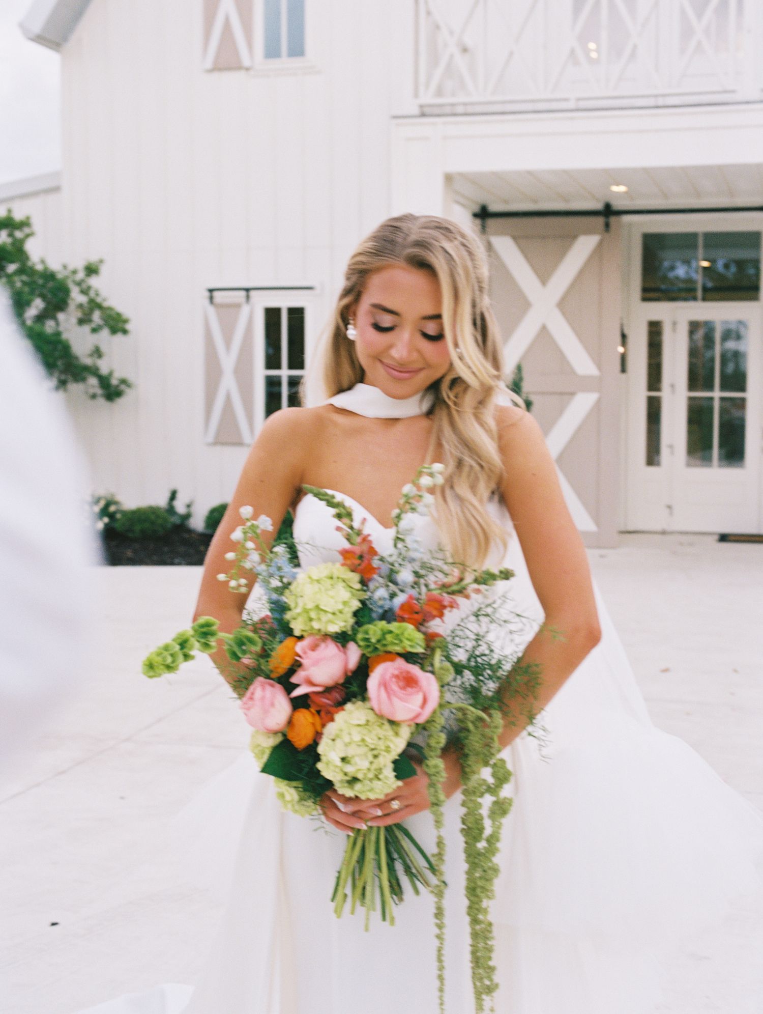 Bride in strapless white gown holding colorful bouquet with pink roses and green hydrangeas outside white barn venue.
