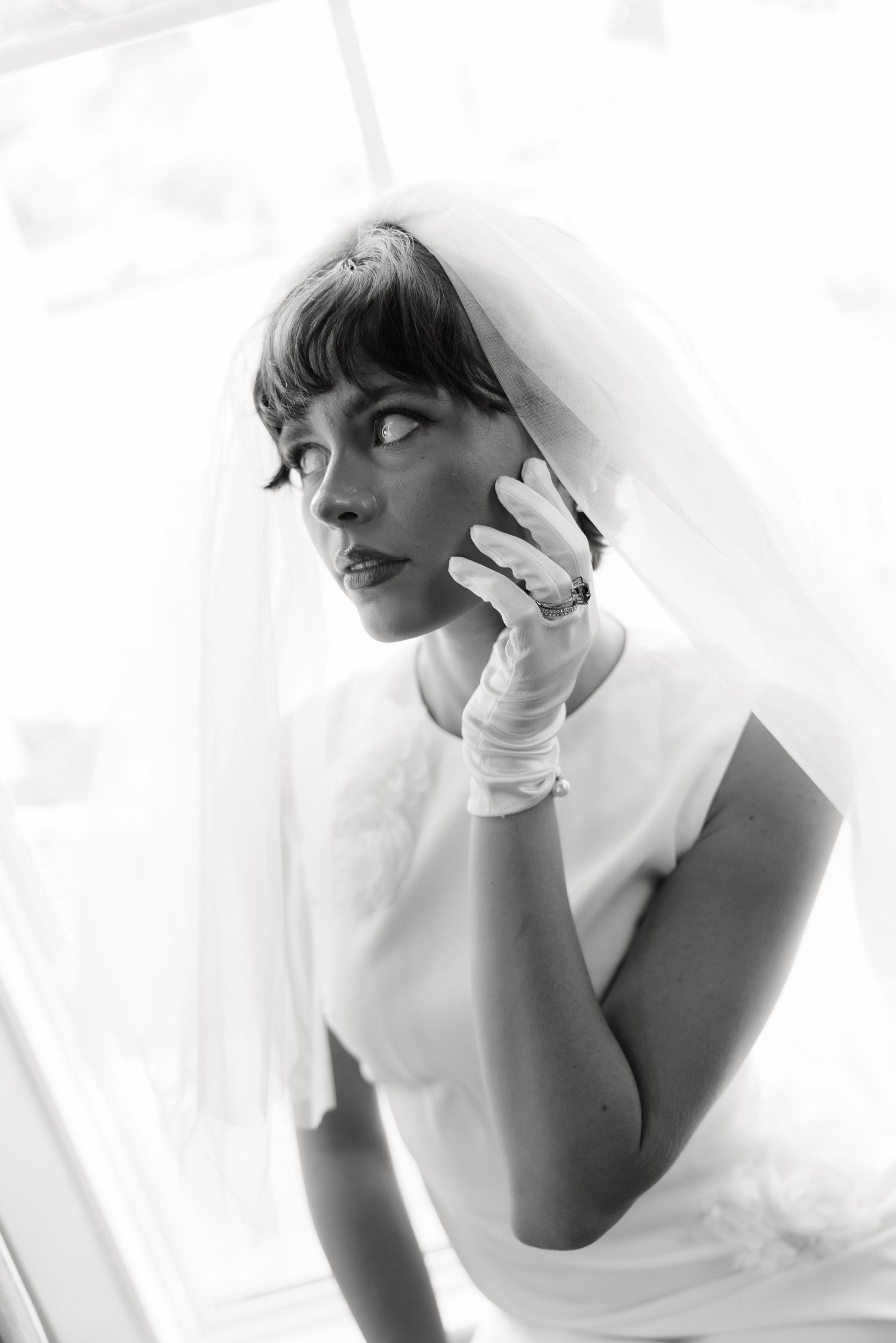 Black and white portrait of a bride in veil and white gloves gazing sideways near a bright window.